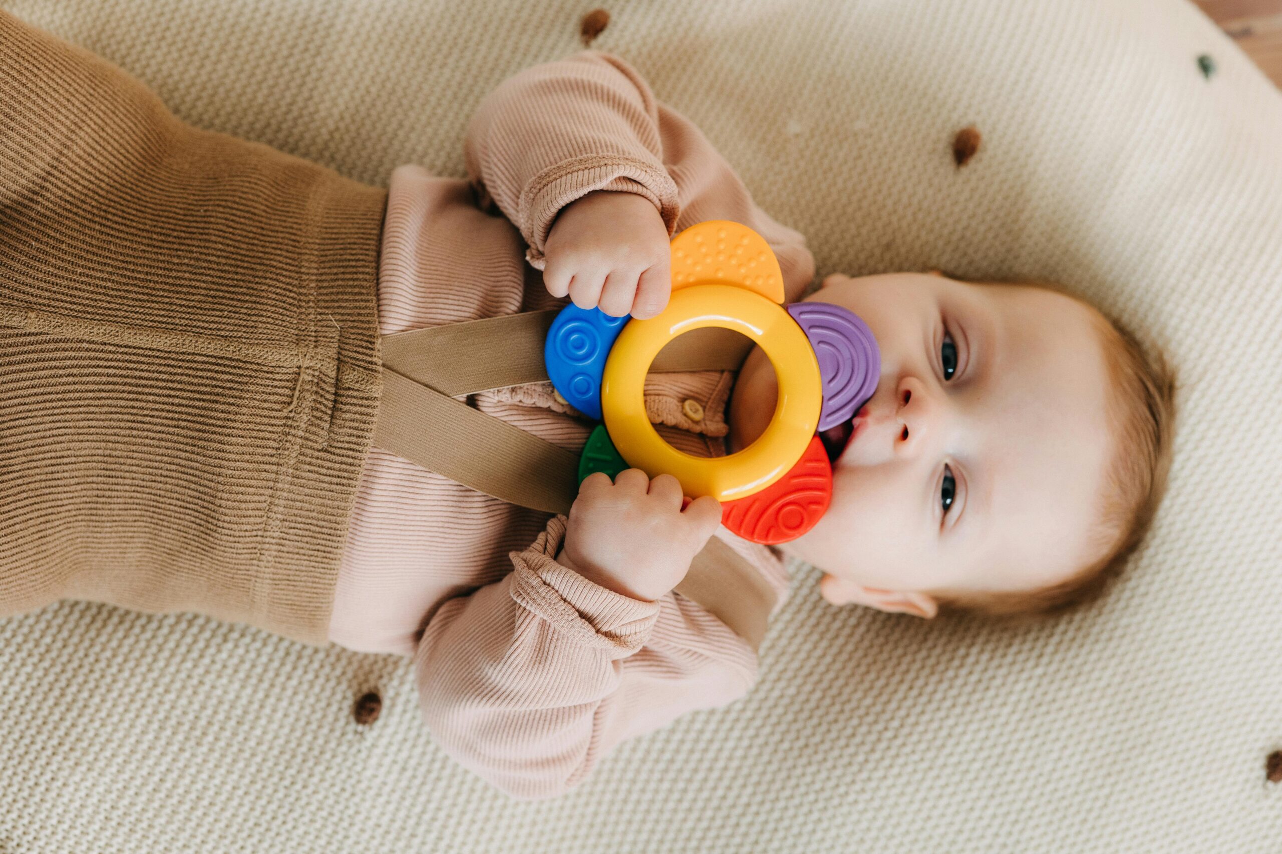 Cute baby chewing on a colorful teething toy while lying on a soft surface.